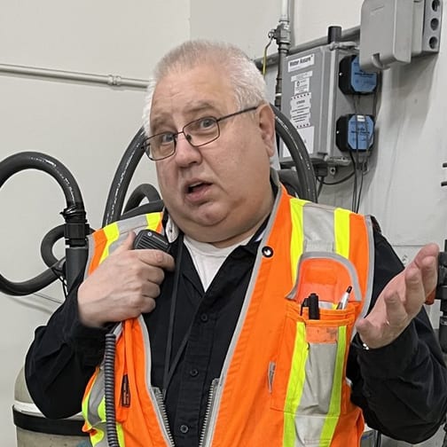 Jay Olivo on a plant floor in a hi-vis vest, holding a two-way radio, talking with one hand raised.